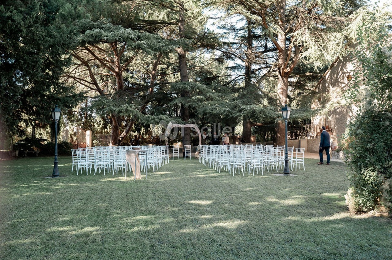 Chaises blanches disposées en rangées sur une pelouse, face à une arche décorative sous de grands arbres.