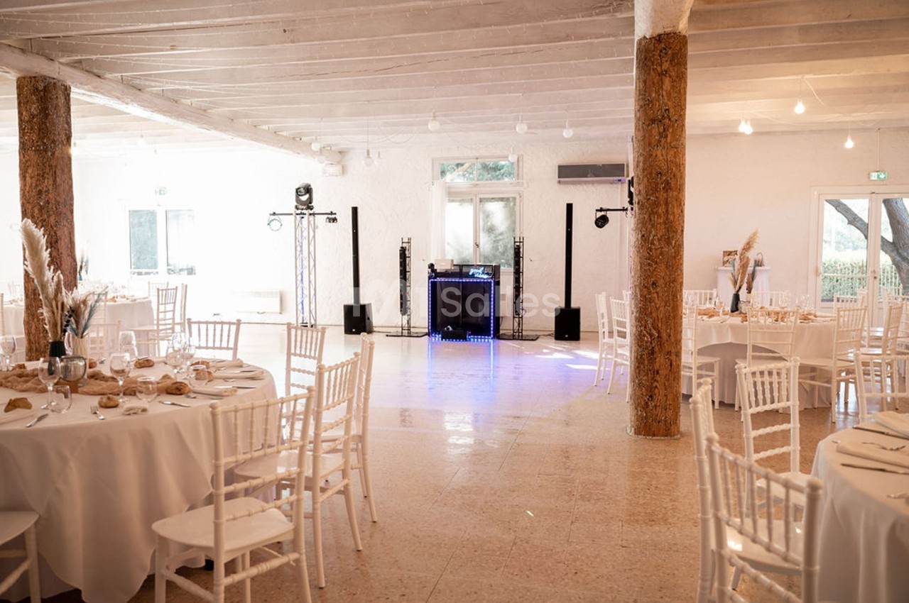 Salle de réception lumineuse avec tables rondes dressées, chaises blanches et espace central dégagé pour une piste de danse.