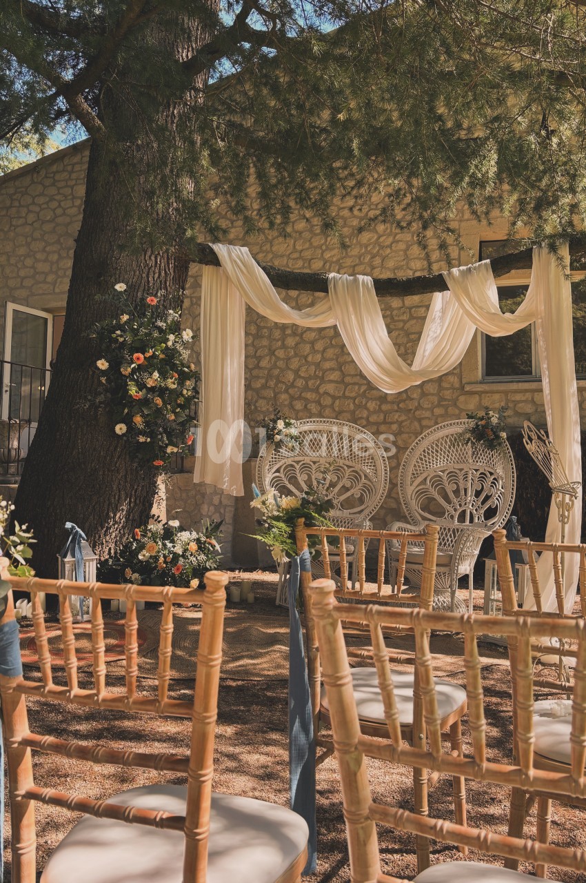 Chaises en bois alignées devant un décor de cérémonie en plein air avec fauteuils en osier, fleurs et voilages suspendus.
