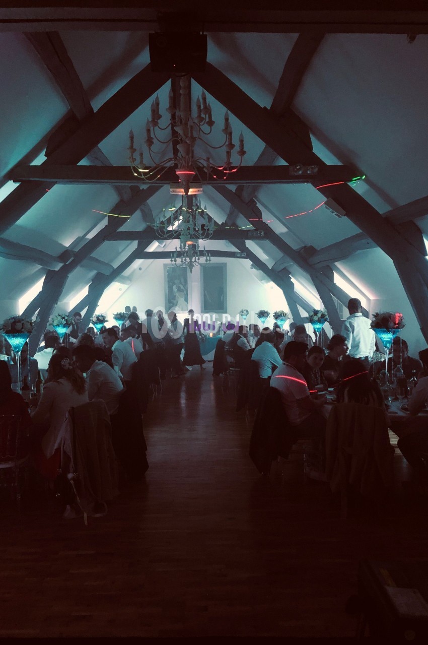 Salle de réception avec des invités assis à des tables décorées, éclairage tamisé et ambiance festive.