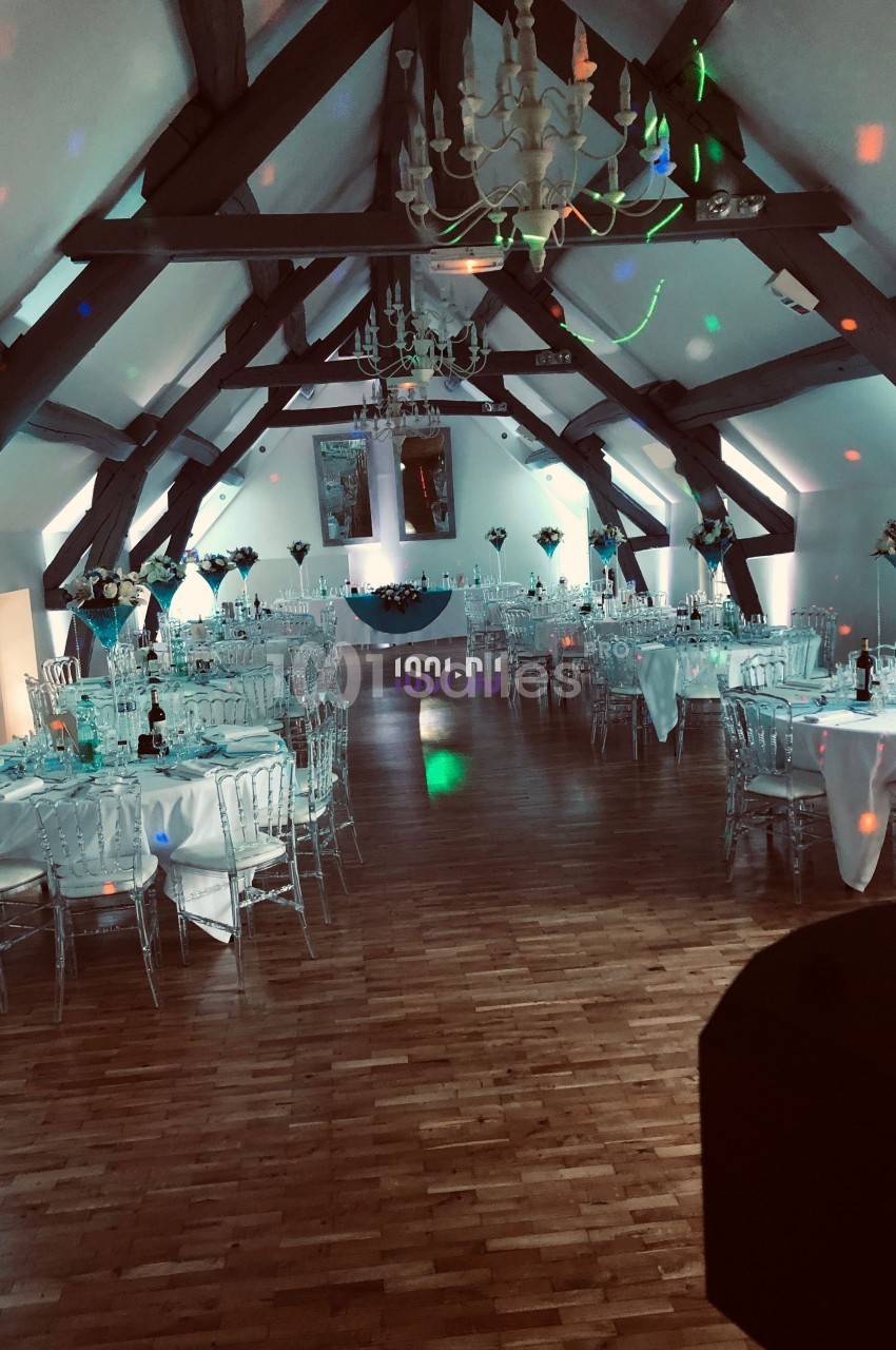 Salle de réception décorée avec des tables dressées, des chaises transparentes et des lumières colorées sur le parquet.