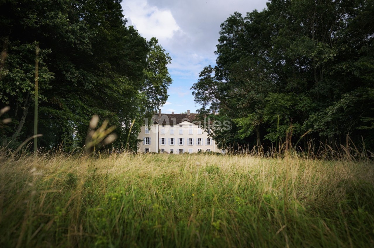 Manoir blanc entouré d'arbres, vu à travers une prairie verdoyante sous un ciel partiellement nuageux.