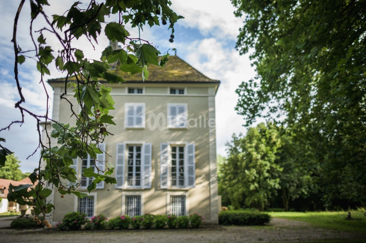 Façade d'une maison ancienne à trois étages, entourée de verdure, sous un ciel partiellement nuageux.