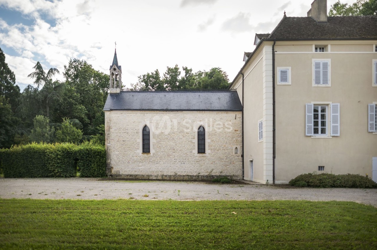 Petite chapelle en pierre attenante à un bâtiment ancien, entourée de verdure et d'un ciel partiellement nuageux.