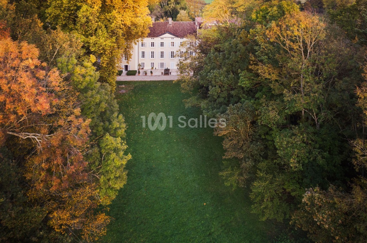Vue aérienne d'un manoir entouré d'arbres aux couleurs automnales, avec une pelouse verdoyante au premier plan.