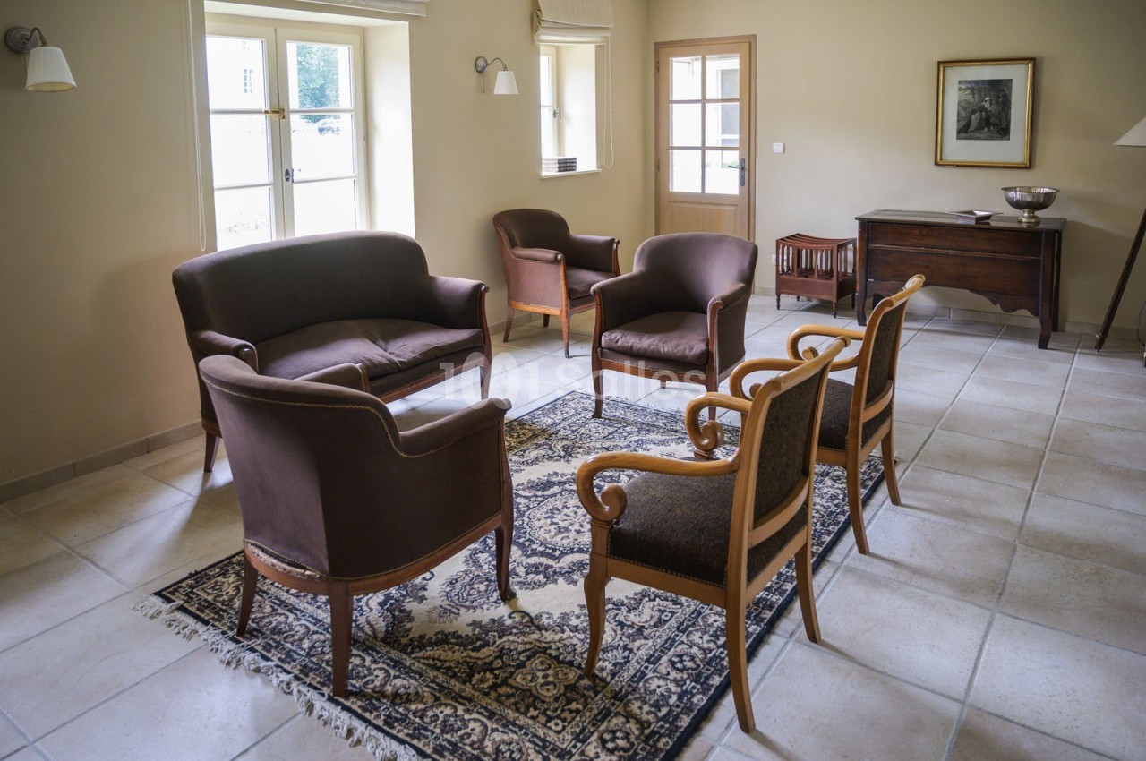 Salon lumineux avec mobilier en bois et tissu brun, tapis à motifs, carrelage clair et fenêtres donnant sur l'extérieur.