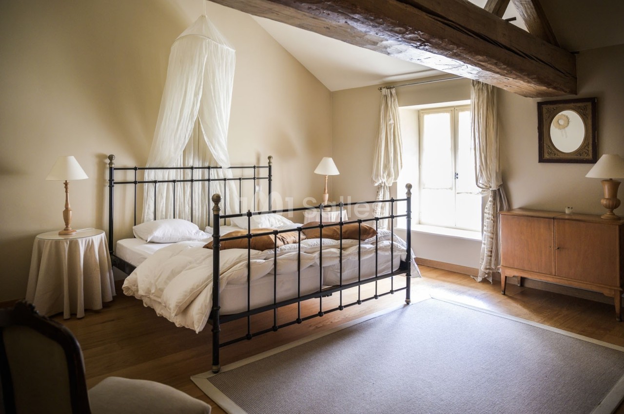 Chambre lumineuse avec lit en métal noir, drap blanc, poutre apparente, commode en bois et grande fenêtre.