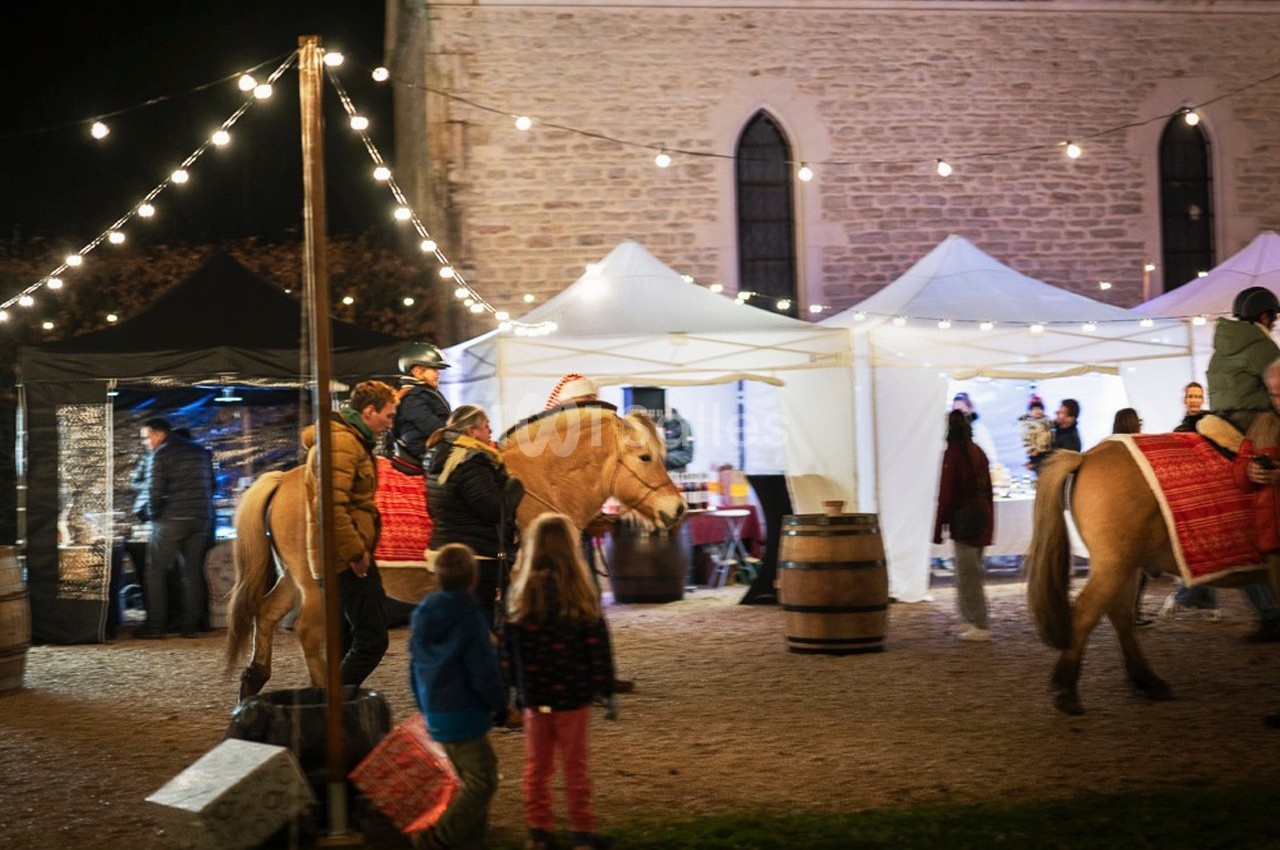 Des chevaux montés par des cavaliers passent devant des stands éclairés lors d'un événement nocturne en plein air.