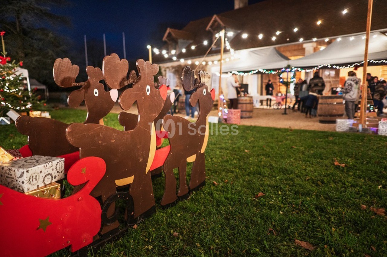 Décoration en bois représentant des rennes et un traîneau, avec des cadeaux, devant un marché de Noël illuminé.