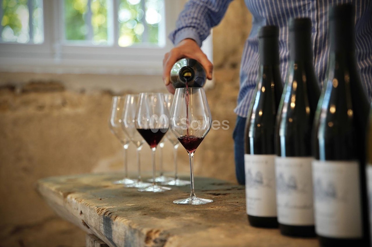Une personne verse du vin rouge dans un verre sur une table en bois, entourée de bouteilles et de verres vides.