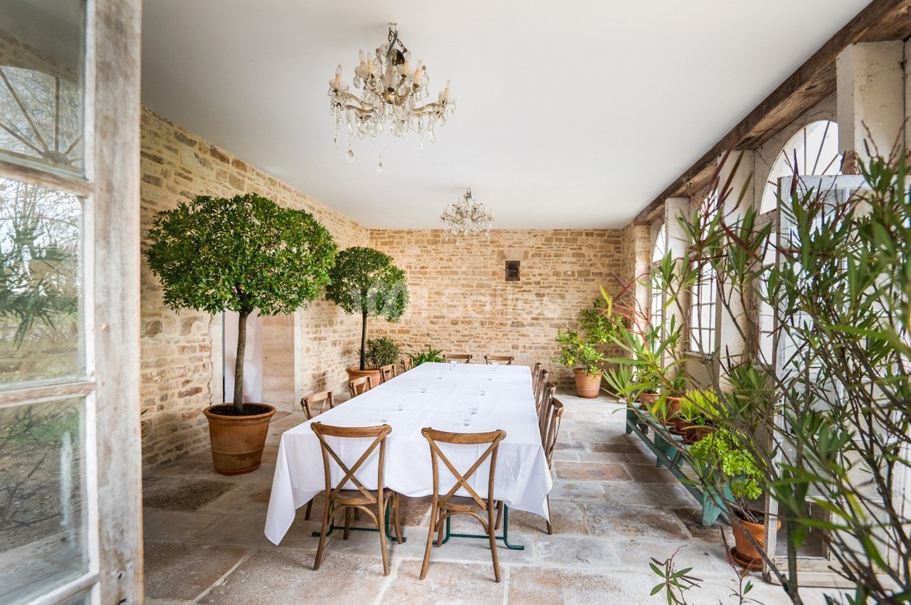 Salle lumineuse avec murs en pierre, table rectangulaire dressée, chaises en bois et plantes en pots.