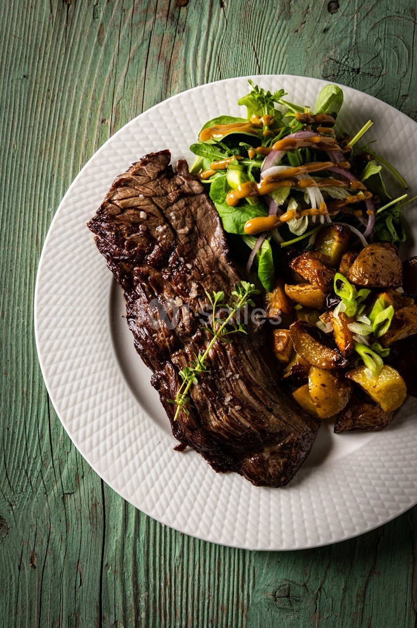 Assiette contenant une pièce de viande grillée, des pommes de terre rôties et une salade verte sur une table en bois.