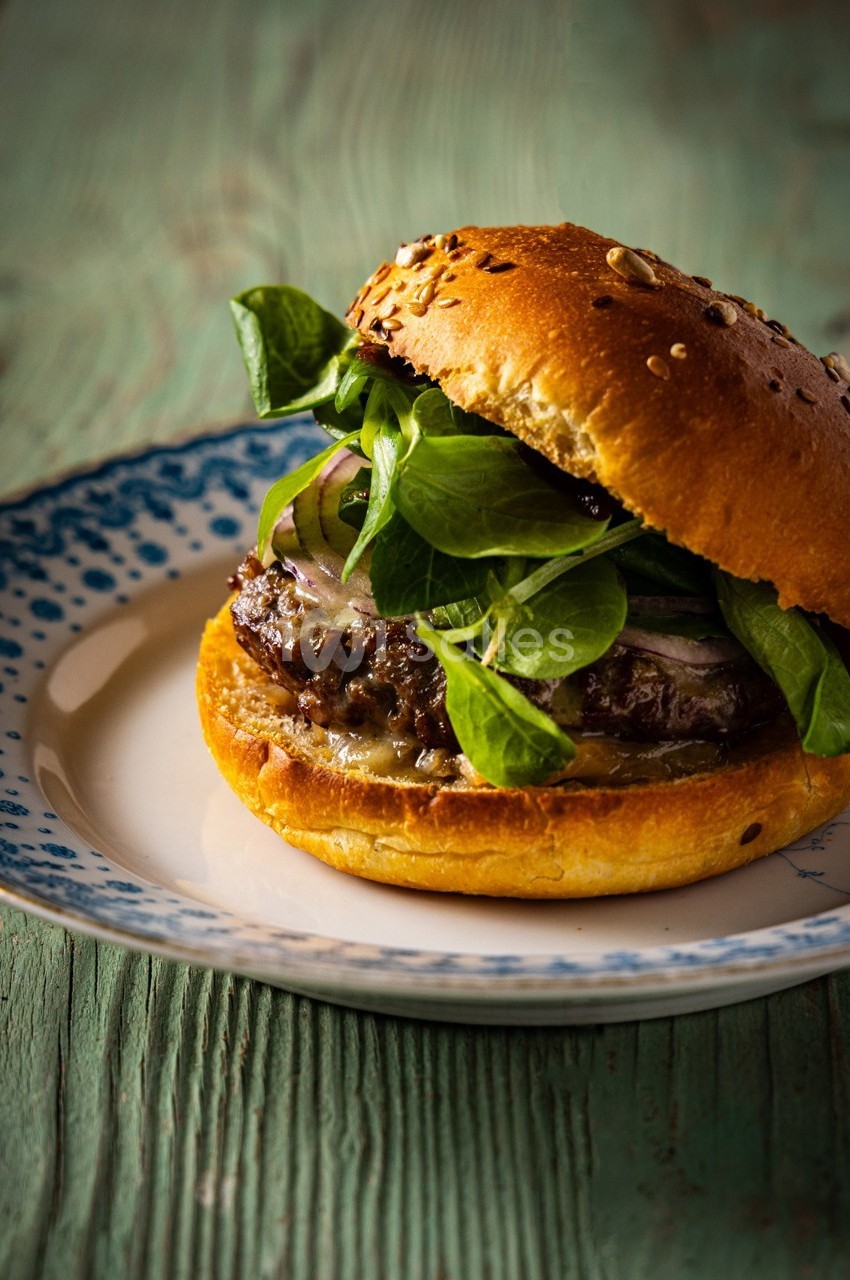 Burger garni de mâche, oignons rouges et fromage fondu, servi dans un pain aux graines sur une assiette blanche et bleue.