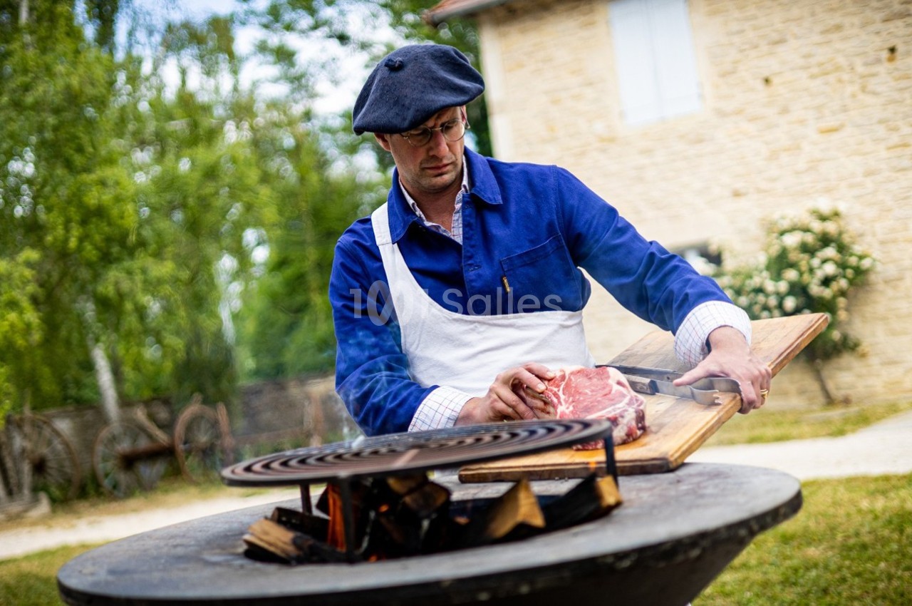 Un homme en tablier et béret prépare un morceau de viande sur une planche près d'un barbecue en plein air.
