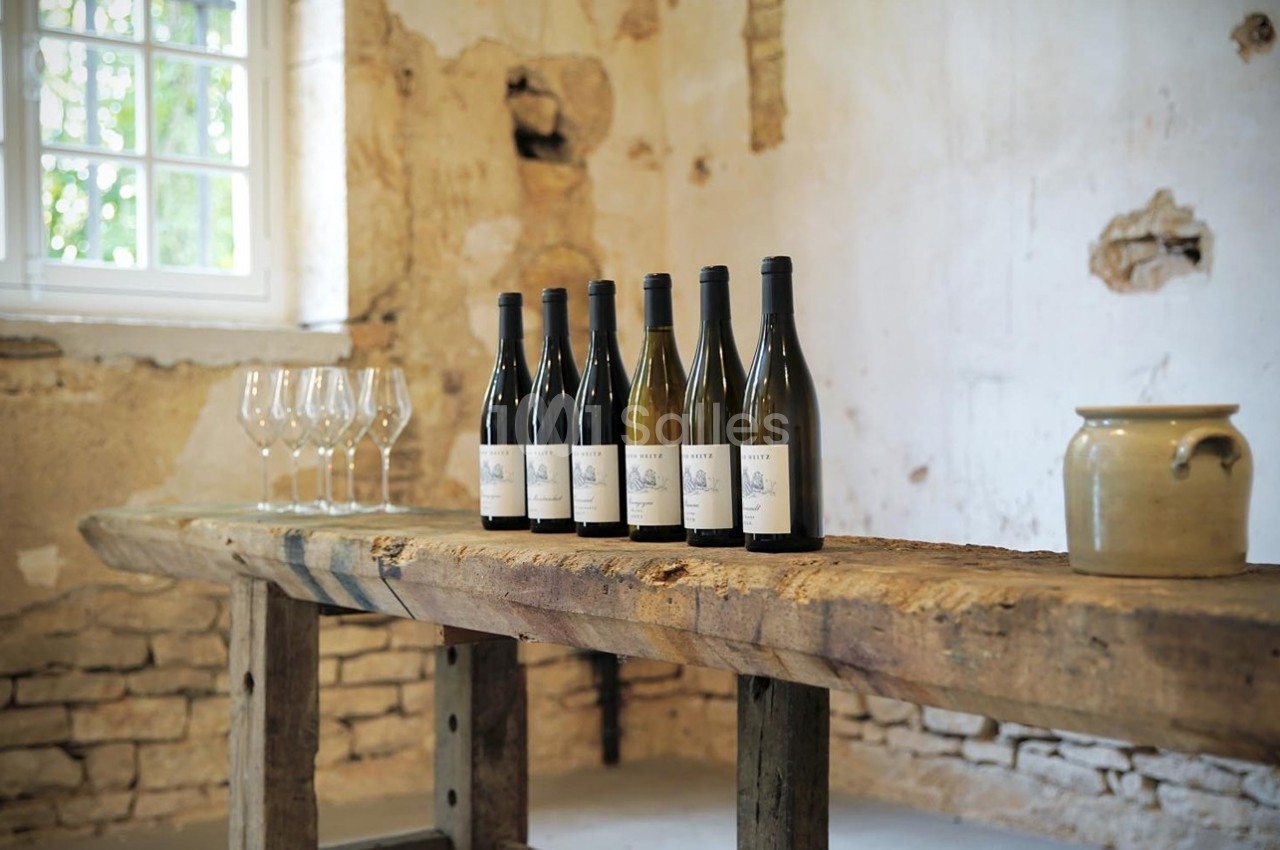 Bouteilles de vin alignées sur une table en bois rustique, avec des verres et un pot en céramique dans une pièce ancienne.