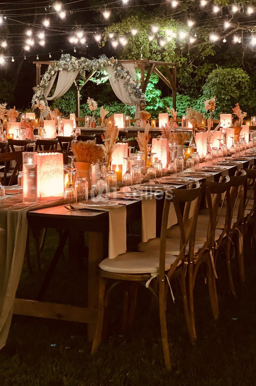 Tables décorées pour un dîner en extérieur, éclairées par des guirlandes lumineuses et ornées de fleurs et bougies.