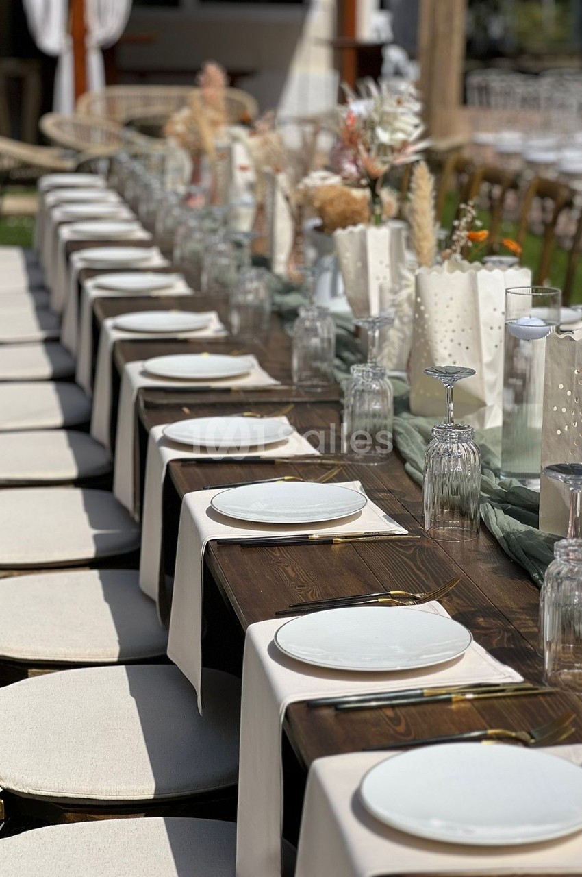 Table en bois dressée pour un repas, avec assiettes blanches, verres et décorations florales dans un cadre extérieur.