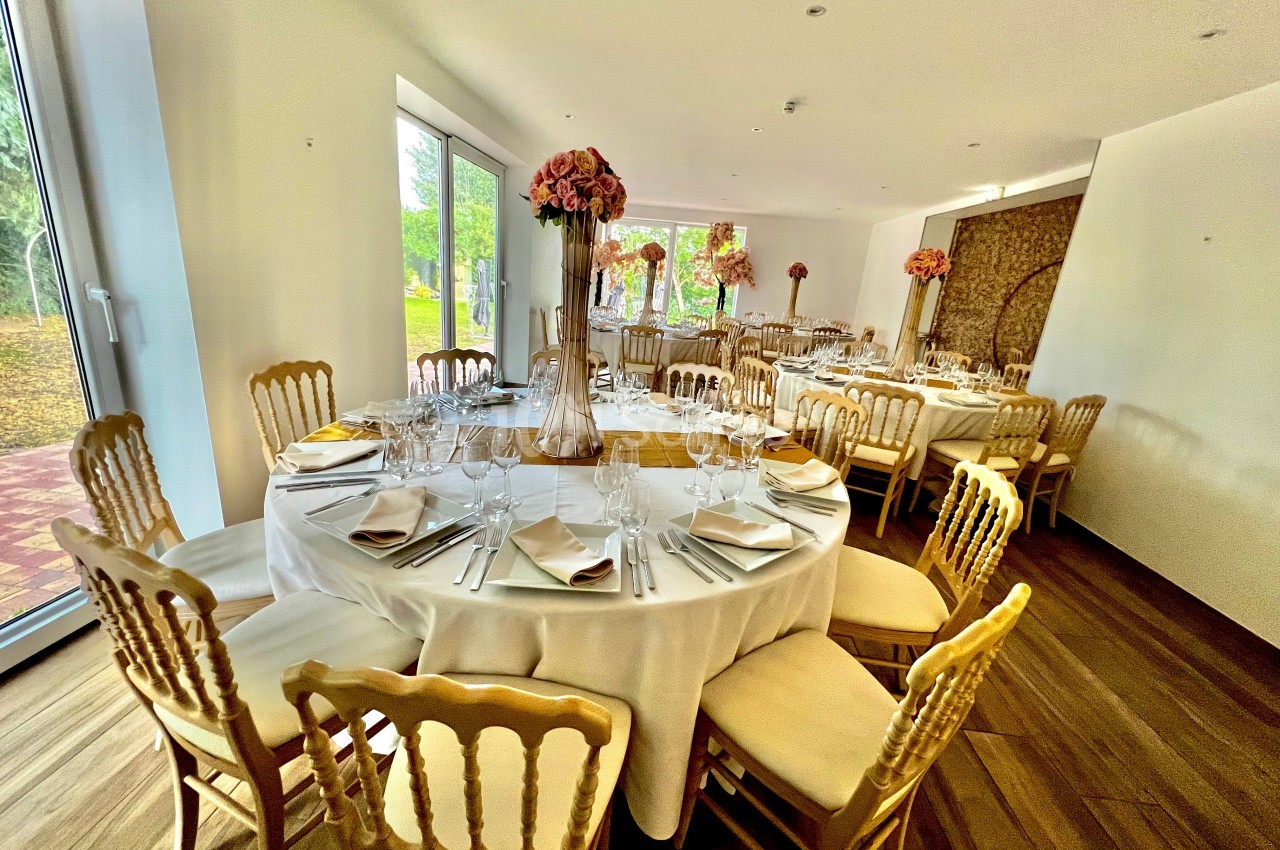 Salle de réception lumineuse avec tables rondes dressées, chaises en bois doré et décorations florales.