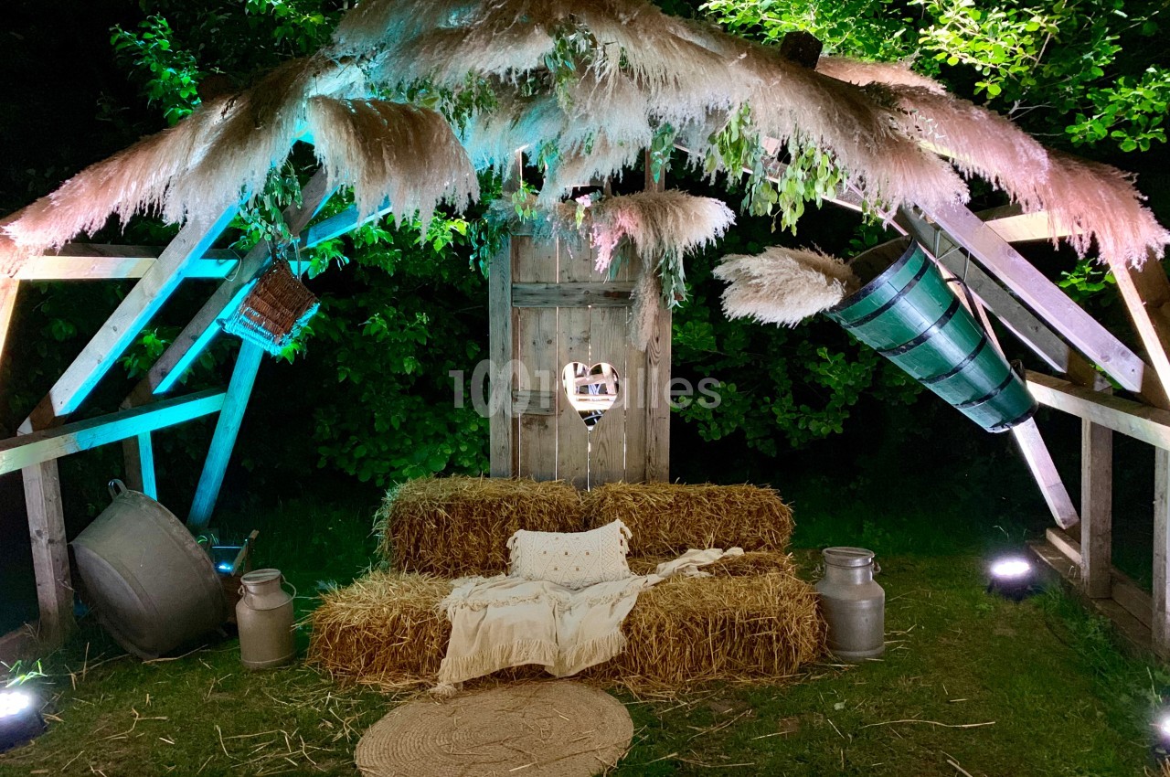 Structure en bois décorée de bottes de paille, de pampas et d'objets rustiques, éclairée par des lumières colorées.