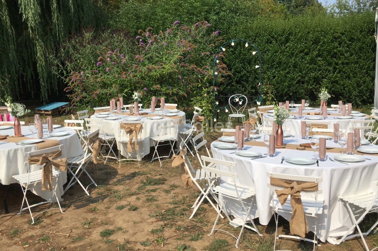 Tables rondes décorées pour un événement en plein air, entourées de chaises blanches et de verdure.