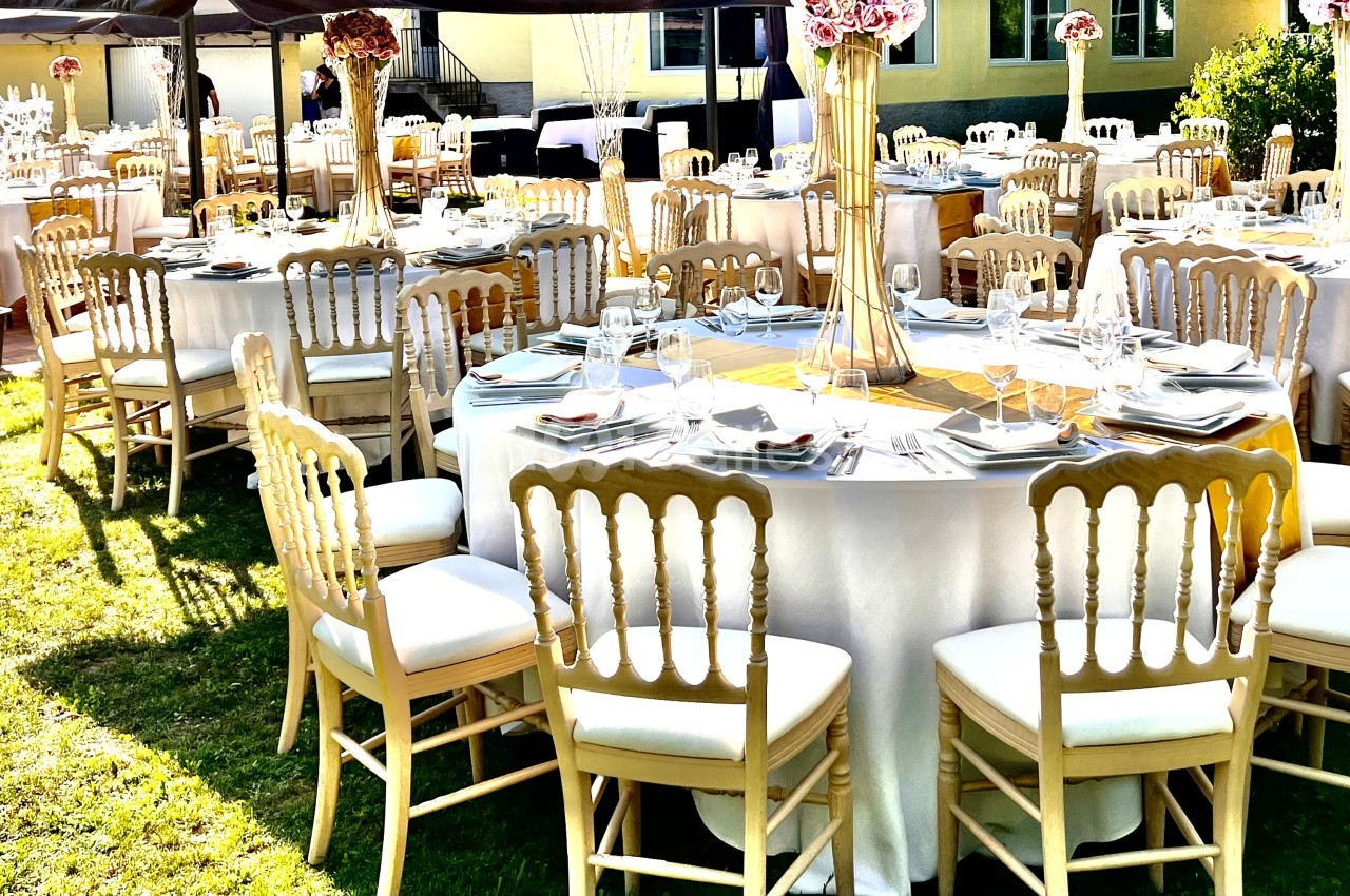 Tables rondes dressées avec nappes blanches, chaises en bois clair et décorations florales dans un jardin ensoleillé.