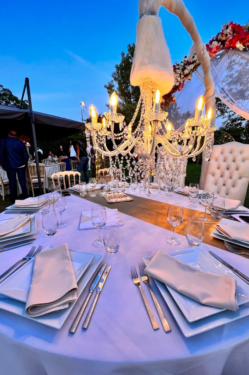 Table élégamment dressée en extérieur avec chandelier lumineux, vaisselle blanche et décor floral en arrière-plan.
