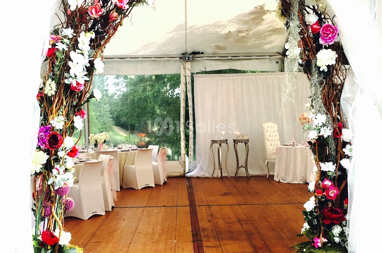 Arche décorée de fleurs colorées dans une tente de réception avec tables et chaises en arrière-plan.