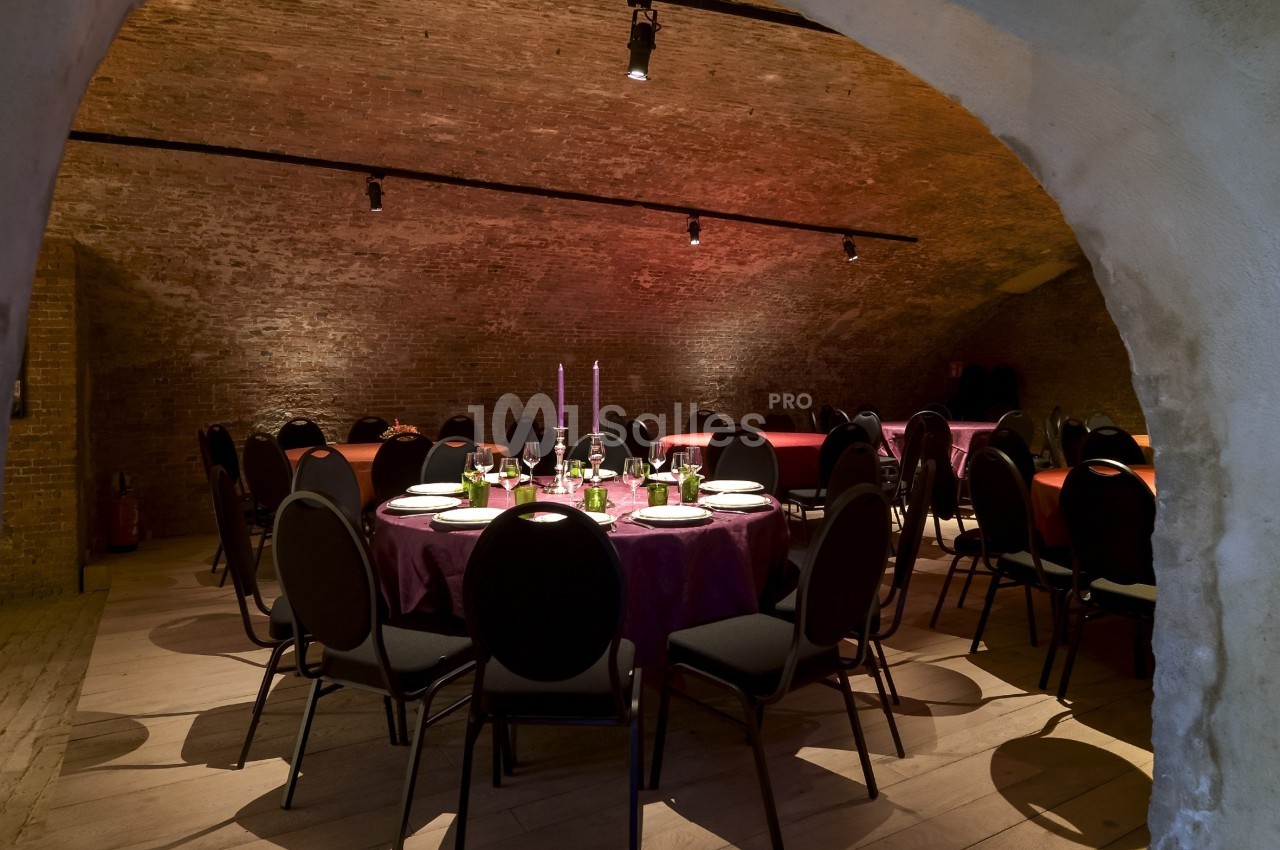 Salle voûtée avec des tables rondes dressées pour un repas, éclairée par des bougies et des lumières tamisées.