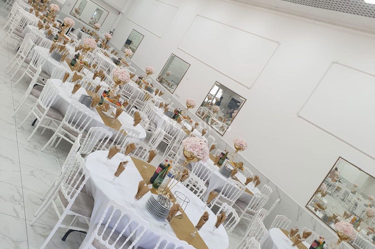 Salle de réception décorée avec des tables rondes, nappes blanches, chaises blanches et centres de table fleuris.