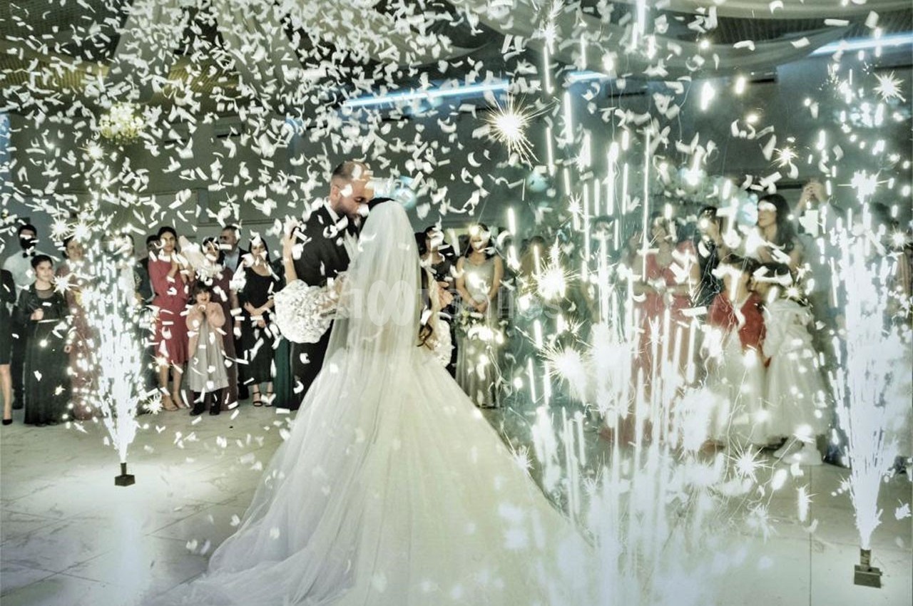 Un couple de mariés danse au centre d'une salle illuminée par des étincelles et des confettis, entouré d'invités.