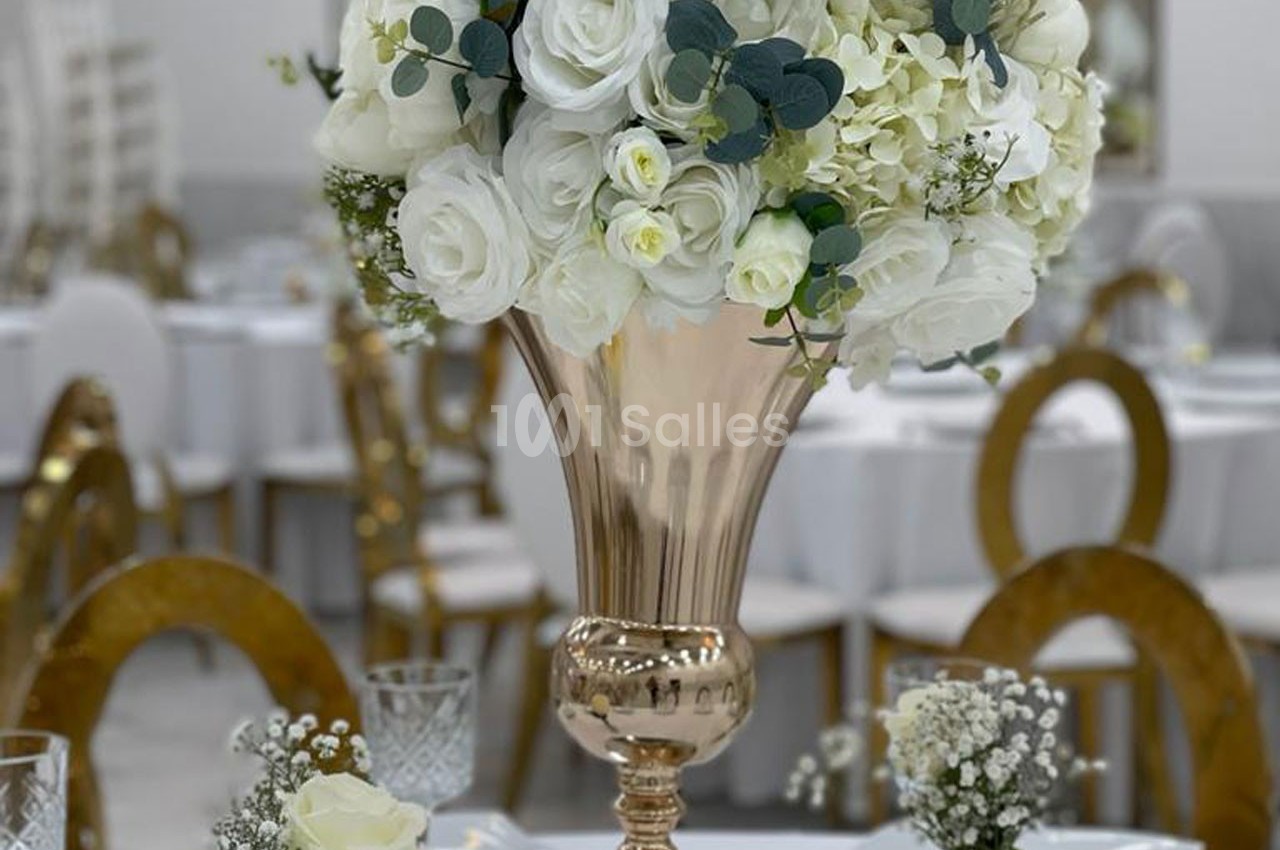 Centre de table avec un vase doré contenant un bouquet de roses blanches et de feuillage, entouré de petites décorations…