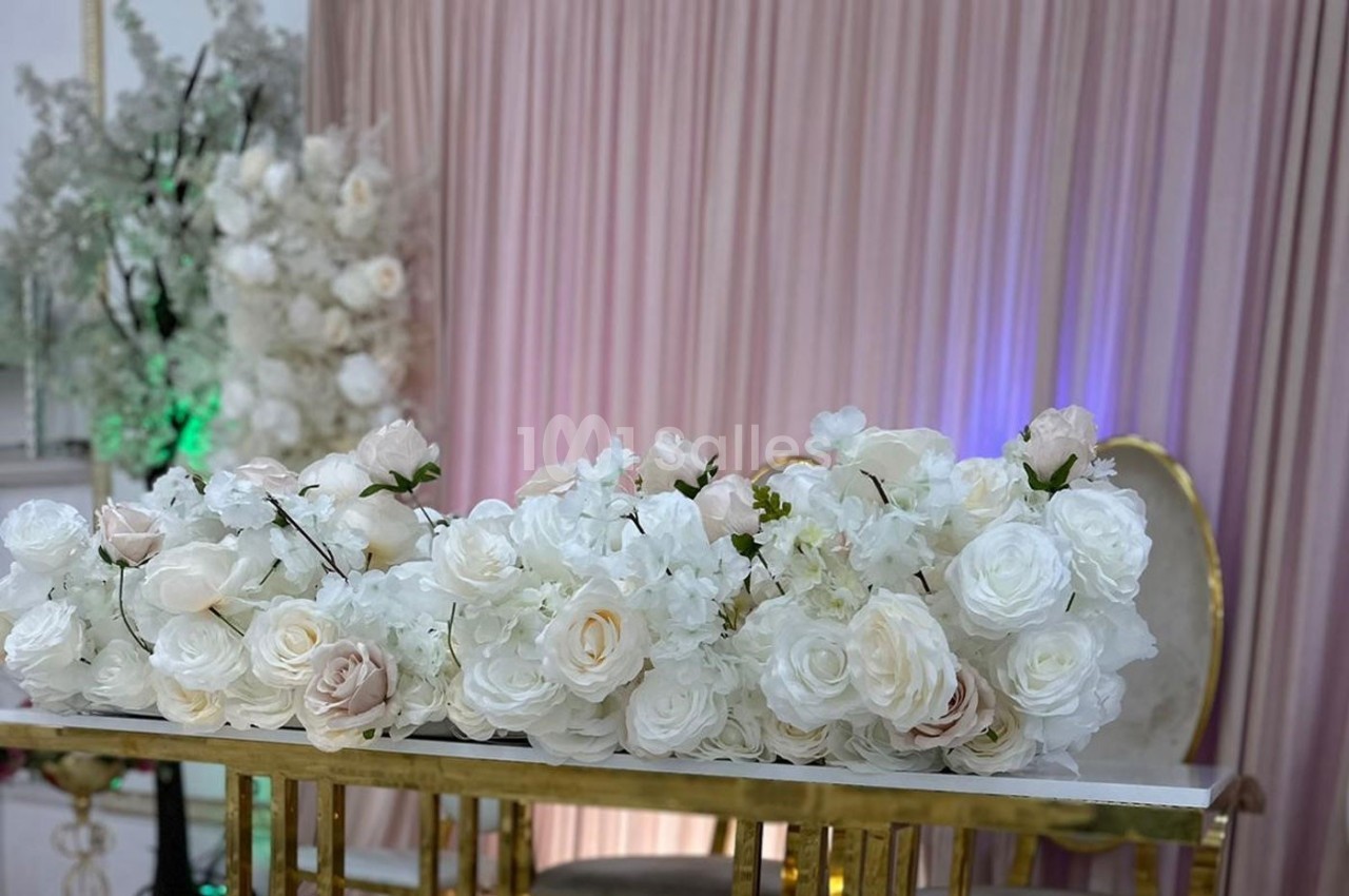 Arrangement floral blanc et rose sur une table dorée, avec un fond de rideaux roses et des décorations florales.