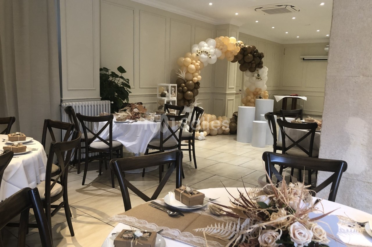 Salle décorée pour un événement avec tables dressées, compositions florales et arche de ballons beige et blanc.