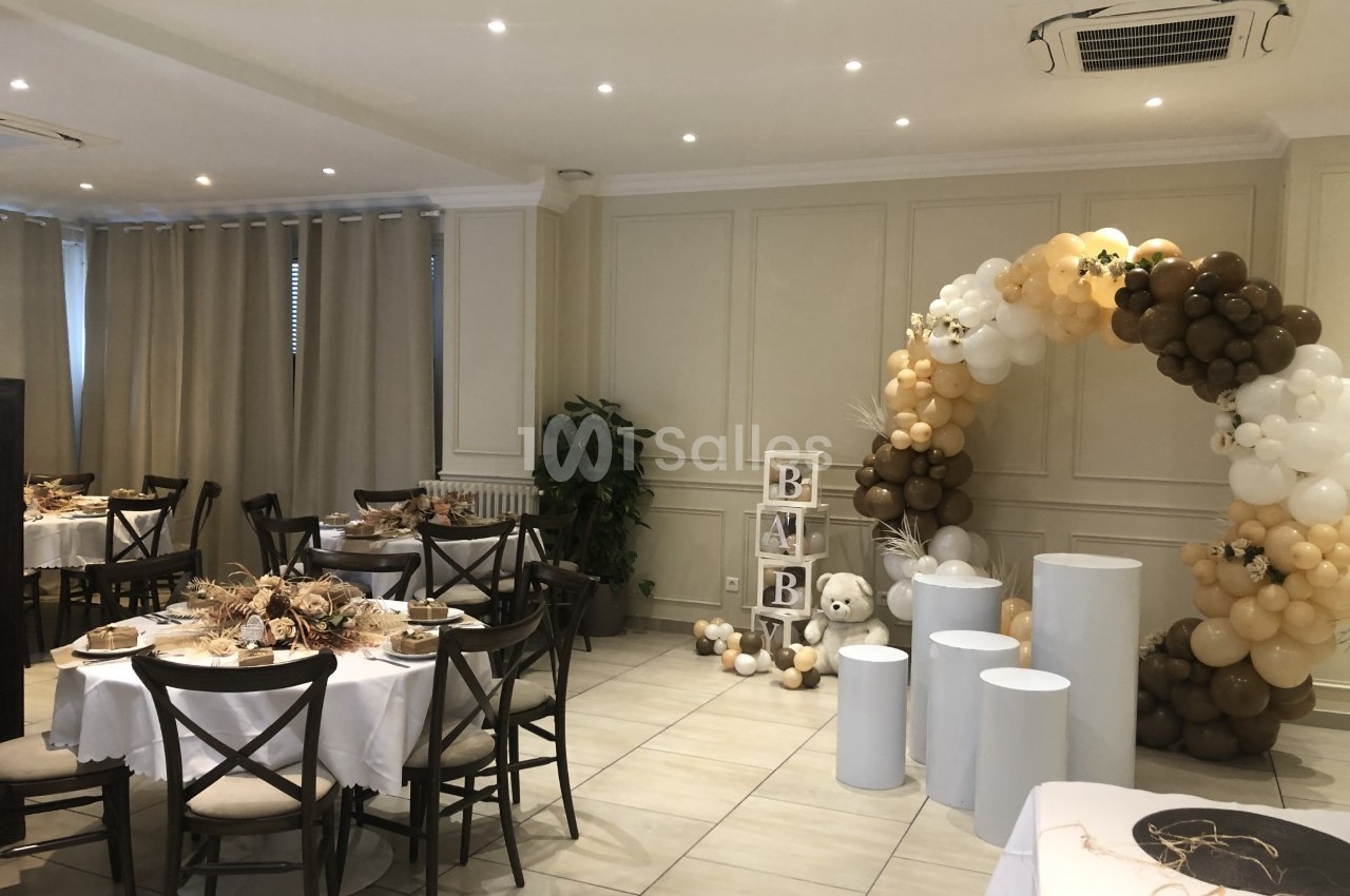Salle décorée pour une fête avec arche de ballons beige et blanc, tables dressées et cubes formant le mot ’BABY’.