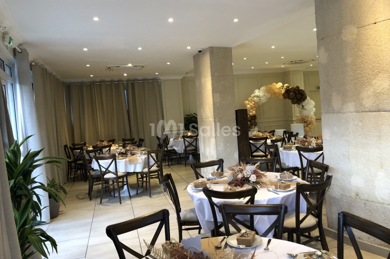 Salle de réception décorée pour un événement, avec tables rondes dressées, chaises en bois et arche florale en arrière-plan.