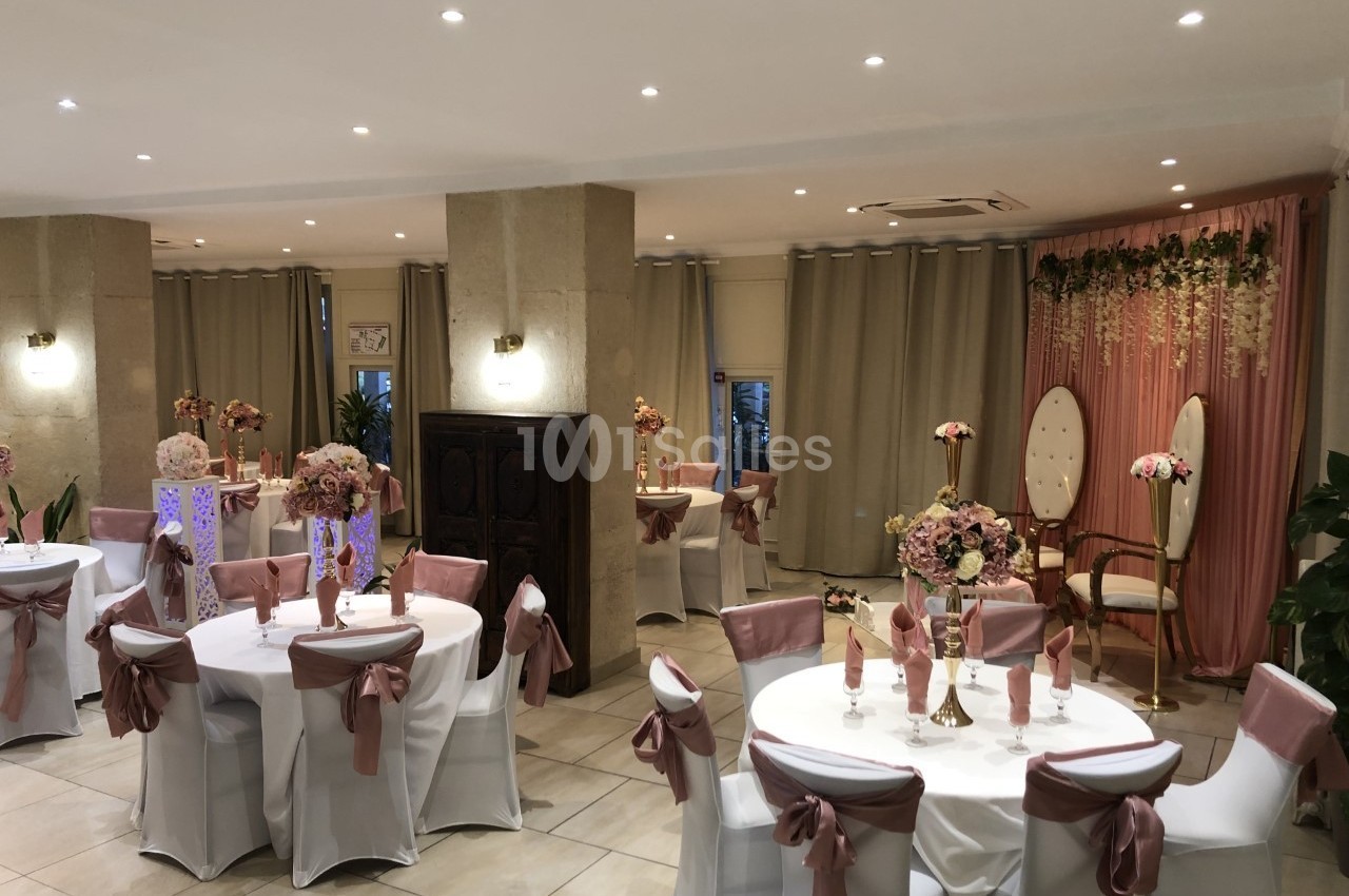Salle de réception décorée avec des tables rondes, nappes blanches, chaises ornées de rubans roses et un fond fleuri.