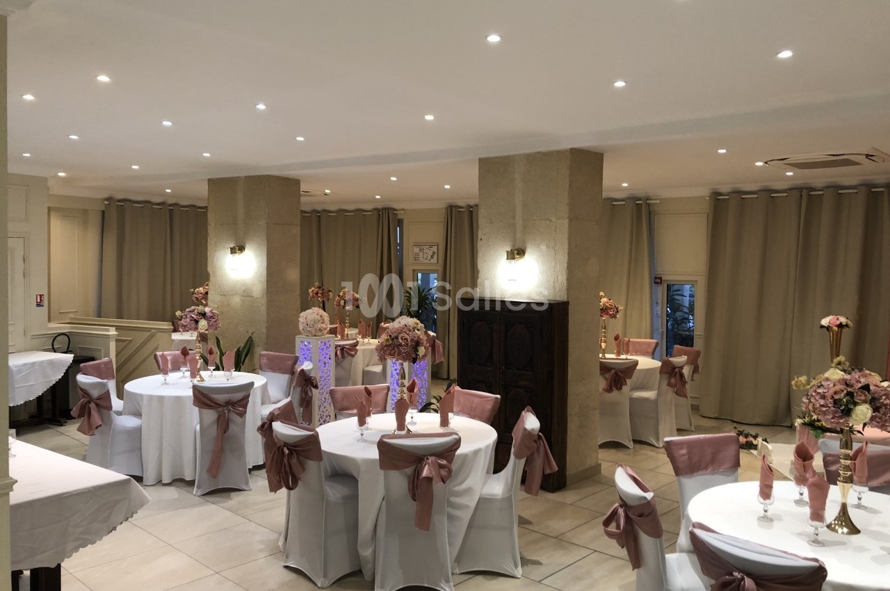 Salle de réception décorée avec des tables rondes, nappes blanches, chaises ornées de nœuds roses et centres de table…