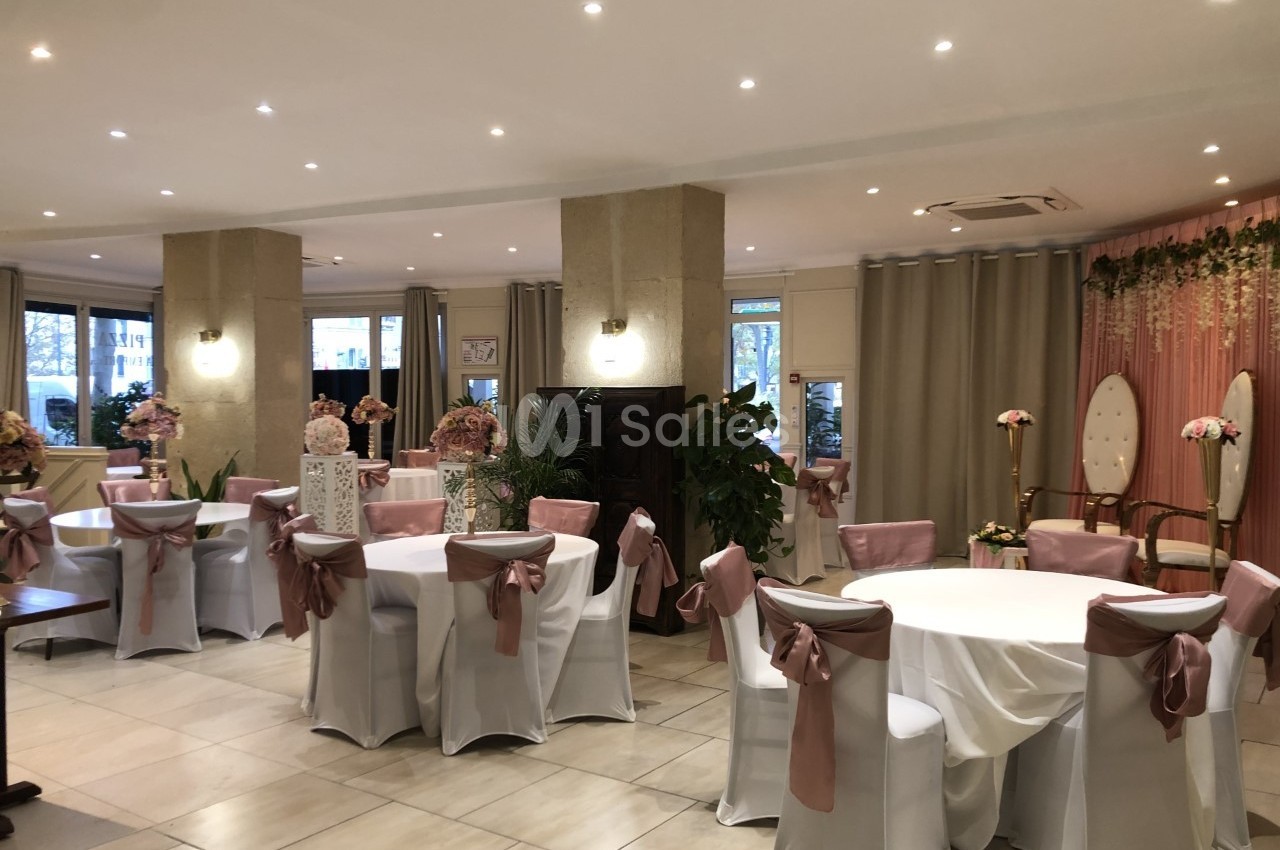 Salle de réception décorée avec des tables rondes, nappes blanches, chaises avec nœuds roses et rideaux beiges.