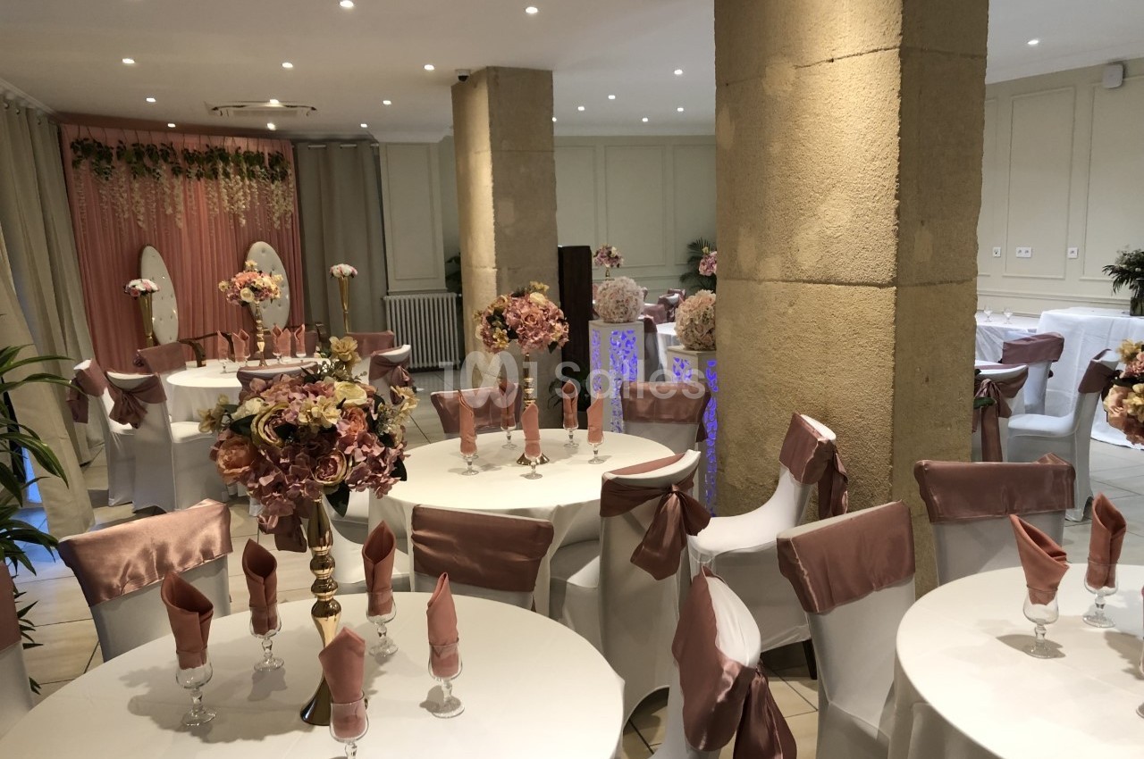 Salle de réception décorée avec des tables rondes, nappes blanches, serviettes roses et compositions florales élégantes.