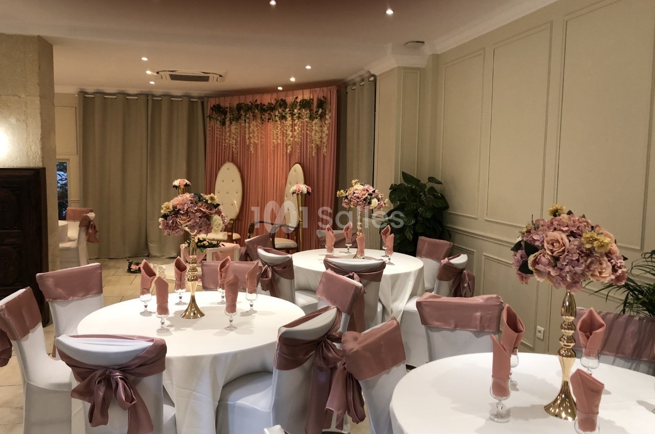 Salle décorée pour un événement avec tables rondes, nappes blanches, chaises ornées de nœuds roses et fleurs en centre de…
