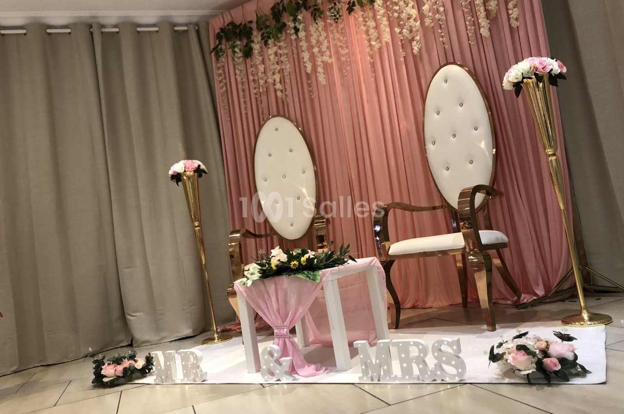Deux fauteuils blancs entourés de décorations florales et d'un rideau rose, disposés pour une cérémonie.
