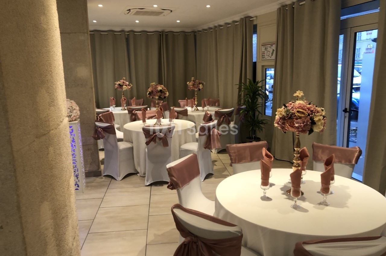 Salle décorée pour un événement avec tables rondes, nappes blanches, chaises ornées de nœuds roses et bouquets floraux.