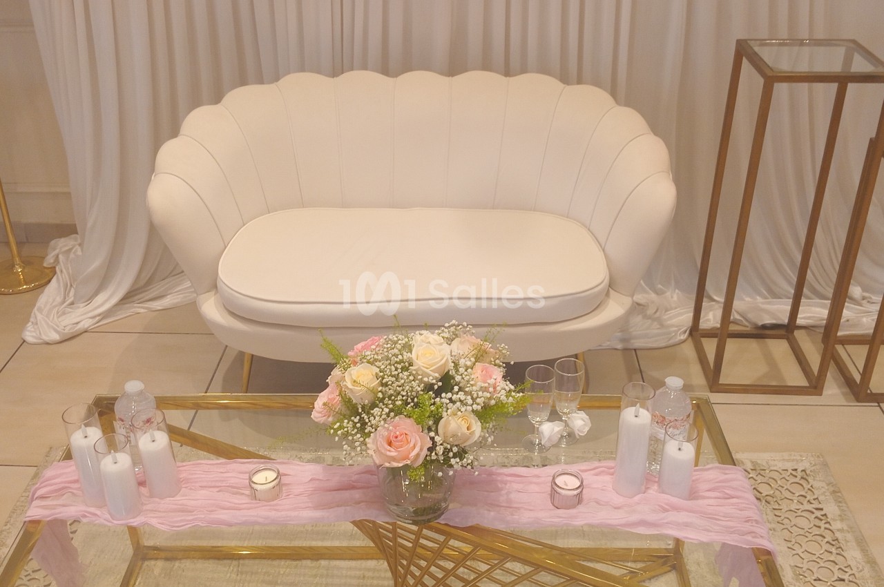 Canapé blanc devant un rideau, table basse dorée décorée de bougies, fleurs et tissu rose.