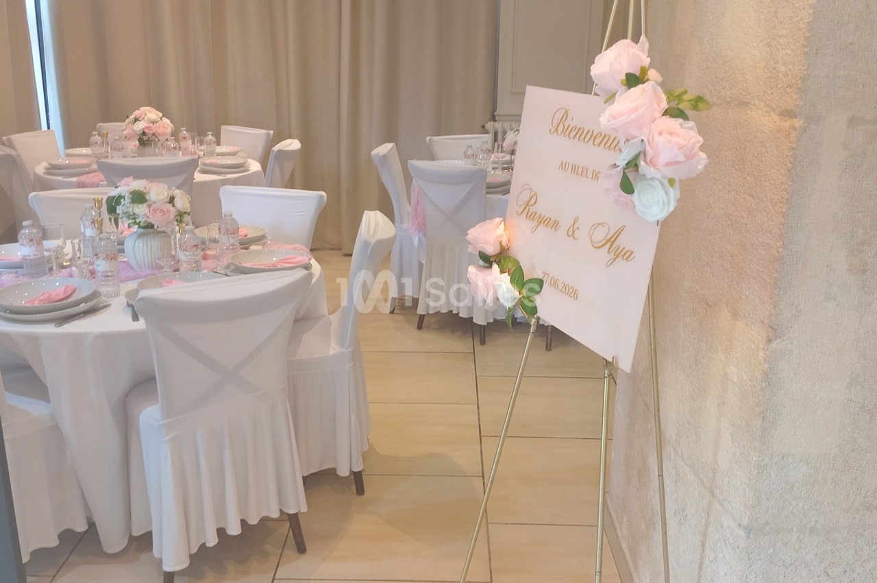 Salle de réception décorée pour un mariage avec des tables dressées et un panneau de bienvenue orné de fleurs.