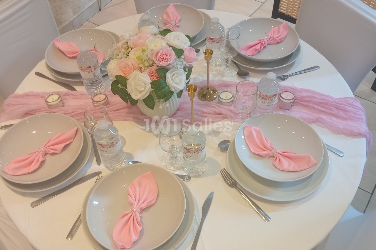 Table ronde élégamment dressée avec nappes blanches, serviettes roses, vaisselle blanche et centre de table floral.