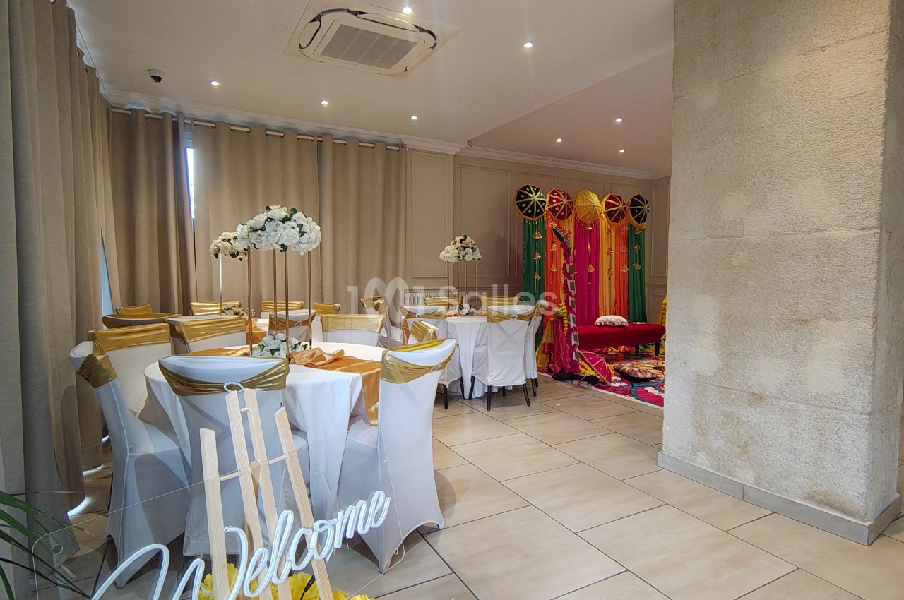 Salle décorée pour un événement, avec chaises blanches ornées de nœuds dorés, fleurs et rideaux colorés en arrière-plan.