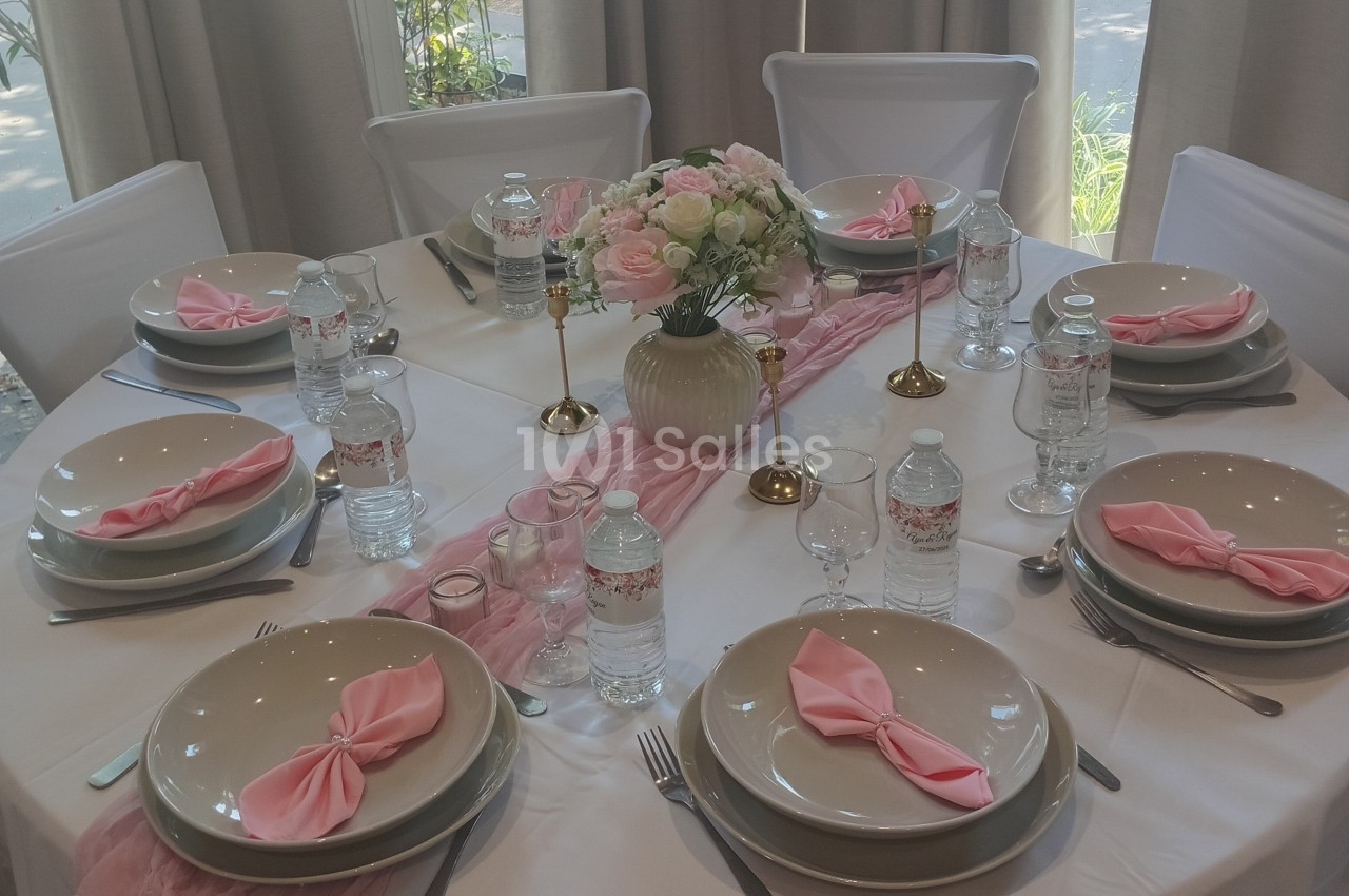 Table élégamment dressée avec vaisselle beige, serviettes roses pliées, bougies dorées et centre de table floral.
