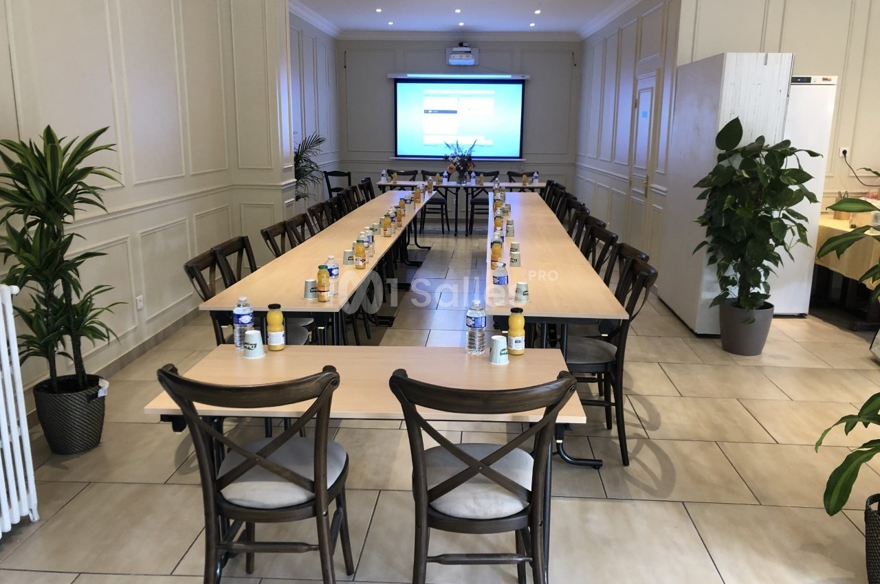 Salle de réunion lumineuse avec tables disposées en U, chaises en bois, écran de projection et plantes décoratives.