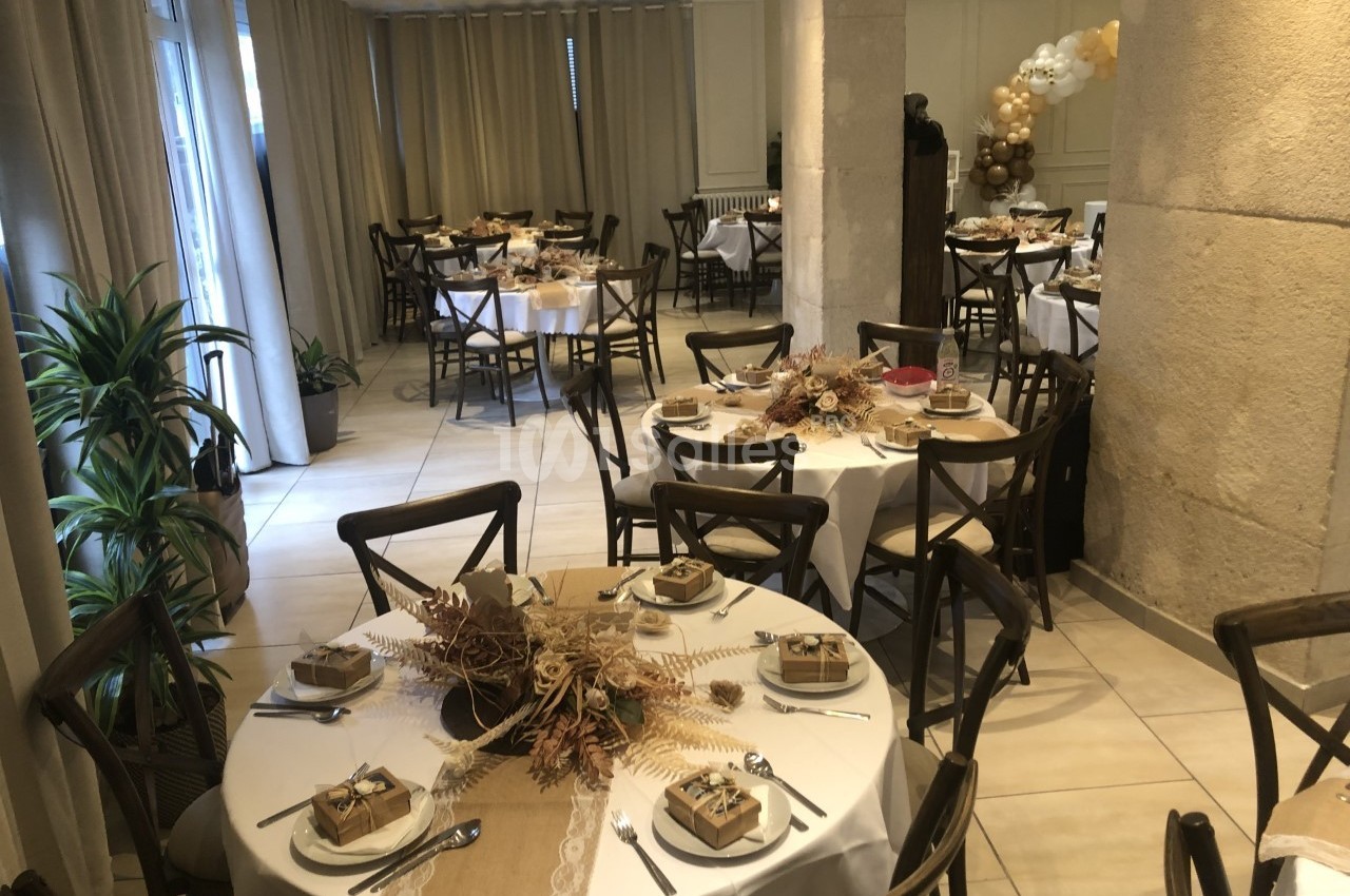 Salle décorée pour un événement, avec des tables rondes dressées, ornées de centres de table naturels et cadeaux.