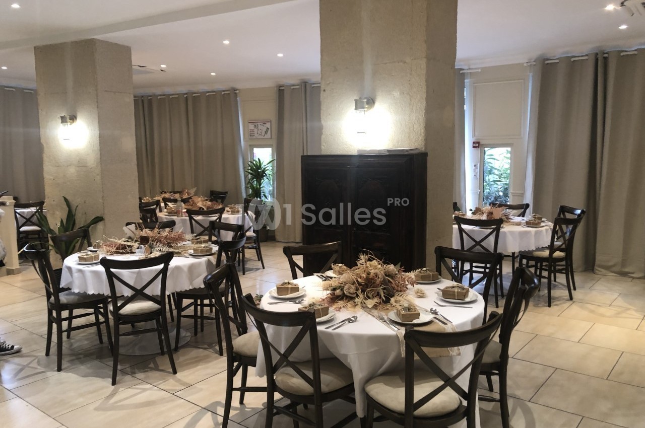 Salle de réception avec tables rondes dressées, décorées de centres de table en fleurs séchées, éclairée par des appliques…